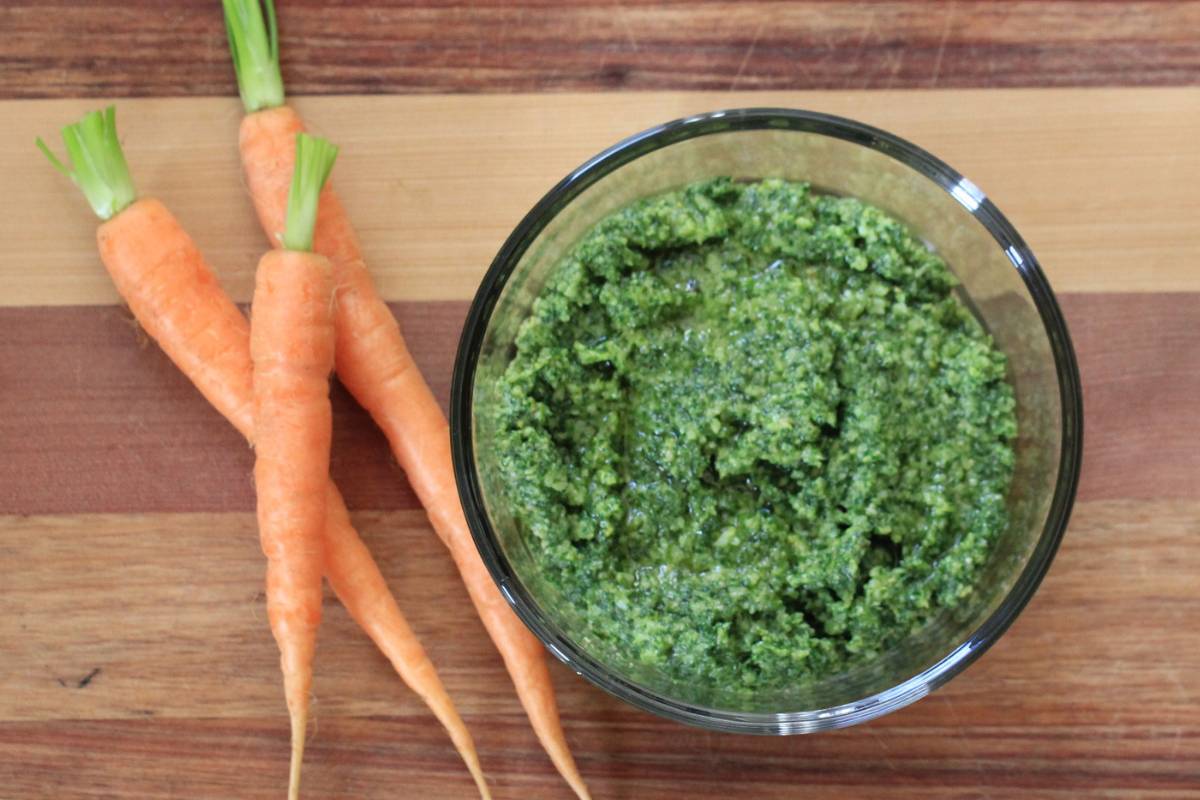 A bowl of carrot top pesto on a wooden chopping board, next to some fresh carrots