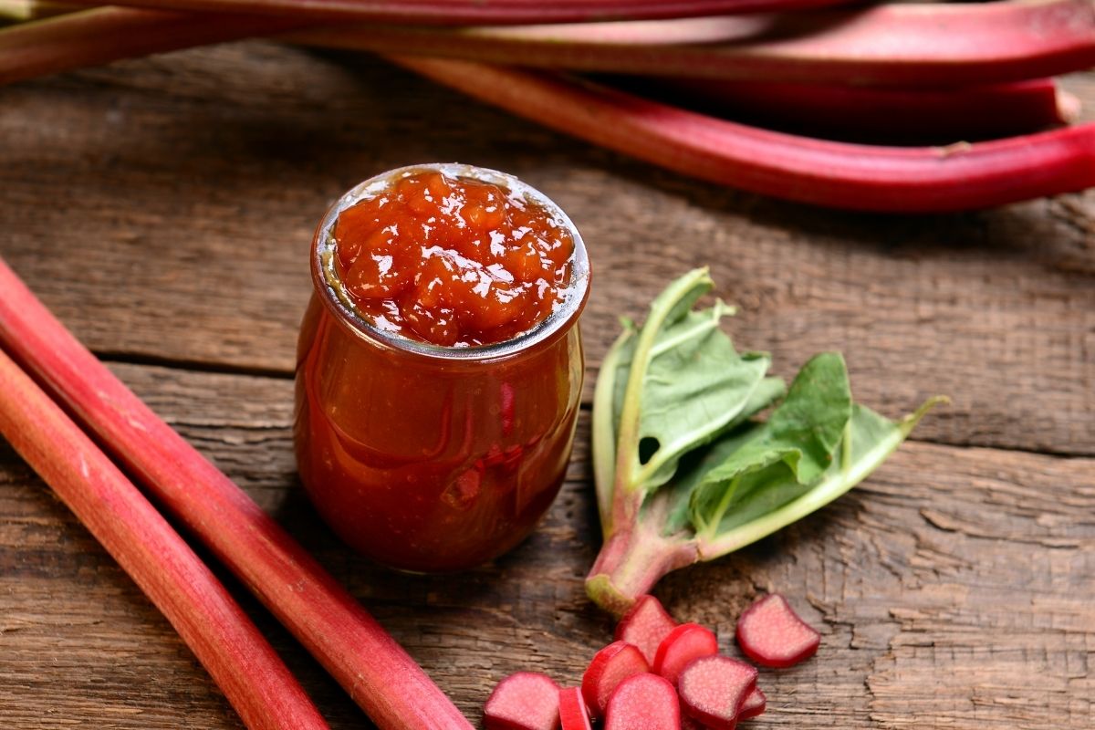 A jar of rhubarb jam with some chopped rhubarb stalks on a kitchen bench