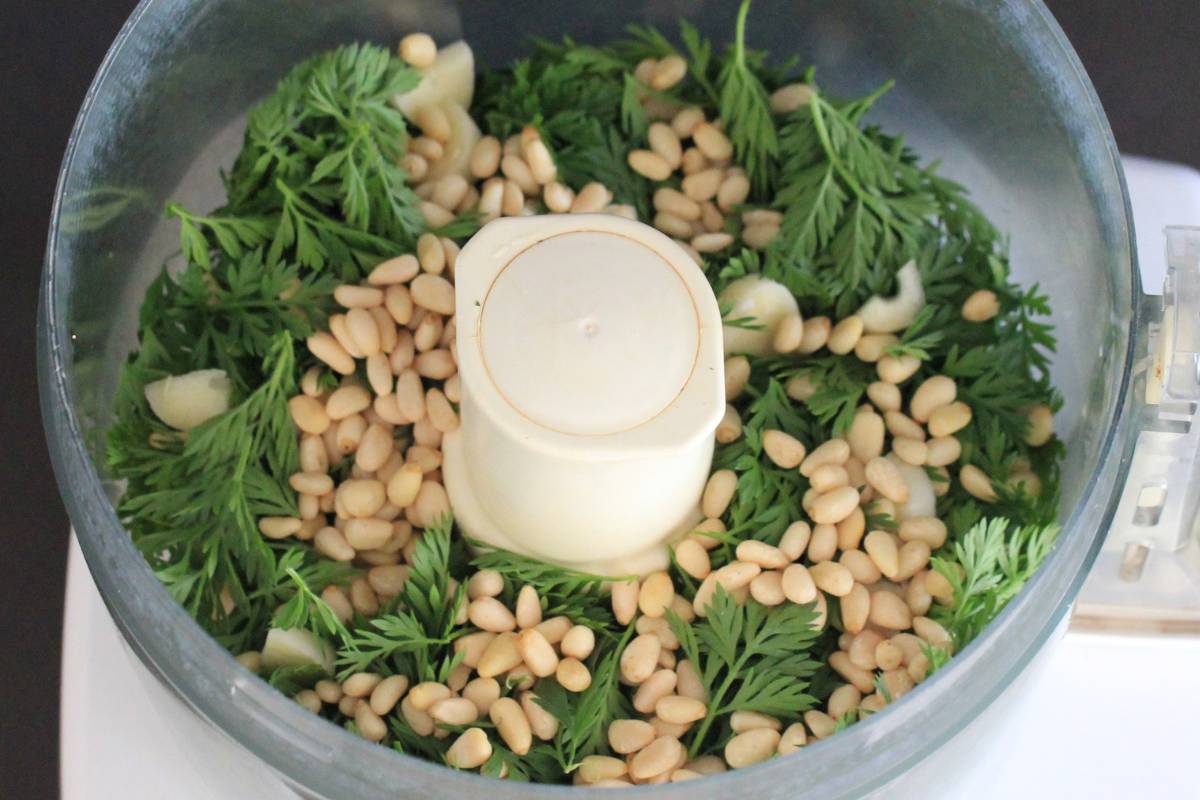 A photo of carrot leaves, garlic cloves and toasted pine nuts in a food processor