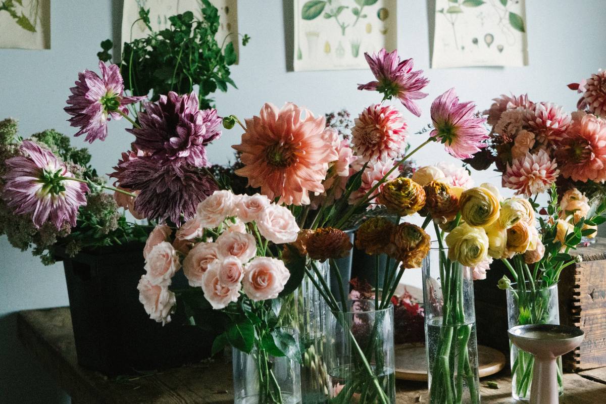 A photo of a row of glass vases filled with flowers on a wooden table in a light-filled room. The flowers are a romantic mix of dahlias, roses and ranunculus.