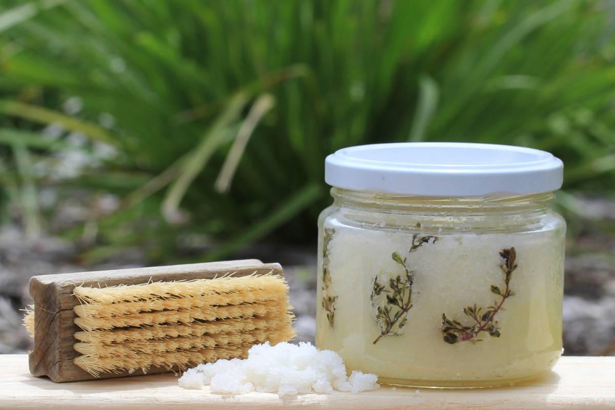 A glass jar of gardener's hand scrub sitting on a wooden board next to a small pile of the scrub and an old nail brush